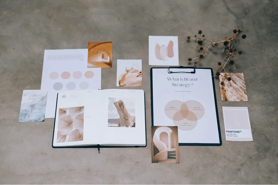 Brand guidelines laid out on a table, including a color palette, a brand strategy diagram on a clipboard, a Pantone swatch and mood board photos in soft neutral tones